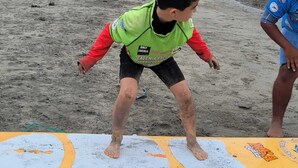A child on a surfboard at a beach with rocky terrain and a building in the background. - Miel de Luna (Cerro Azul)