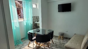A small, modern room with a round glass table and black chairs, a flat-screen TV mounted on the wall, and a cushioned bench. - Miel de Luna (Cerro Azul)