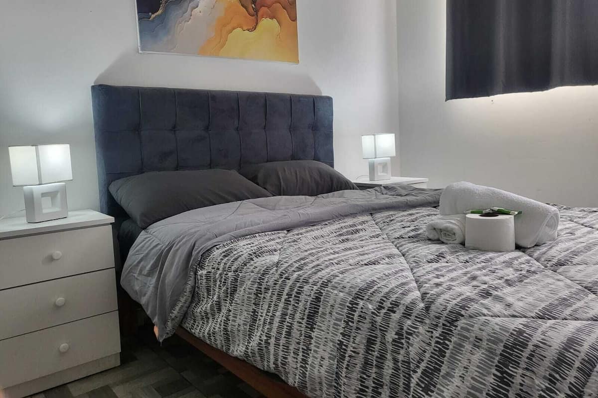 A modern bedroom with a tufted headboard, a grey patterned bedspread, two bedside tables with lamps, and a painting above the bed.