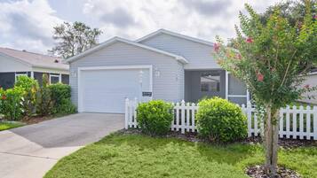 A single-story house with a white garage door, a small front yard with a white picket fence, and a tree with pink flowers.