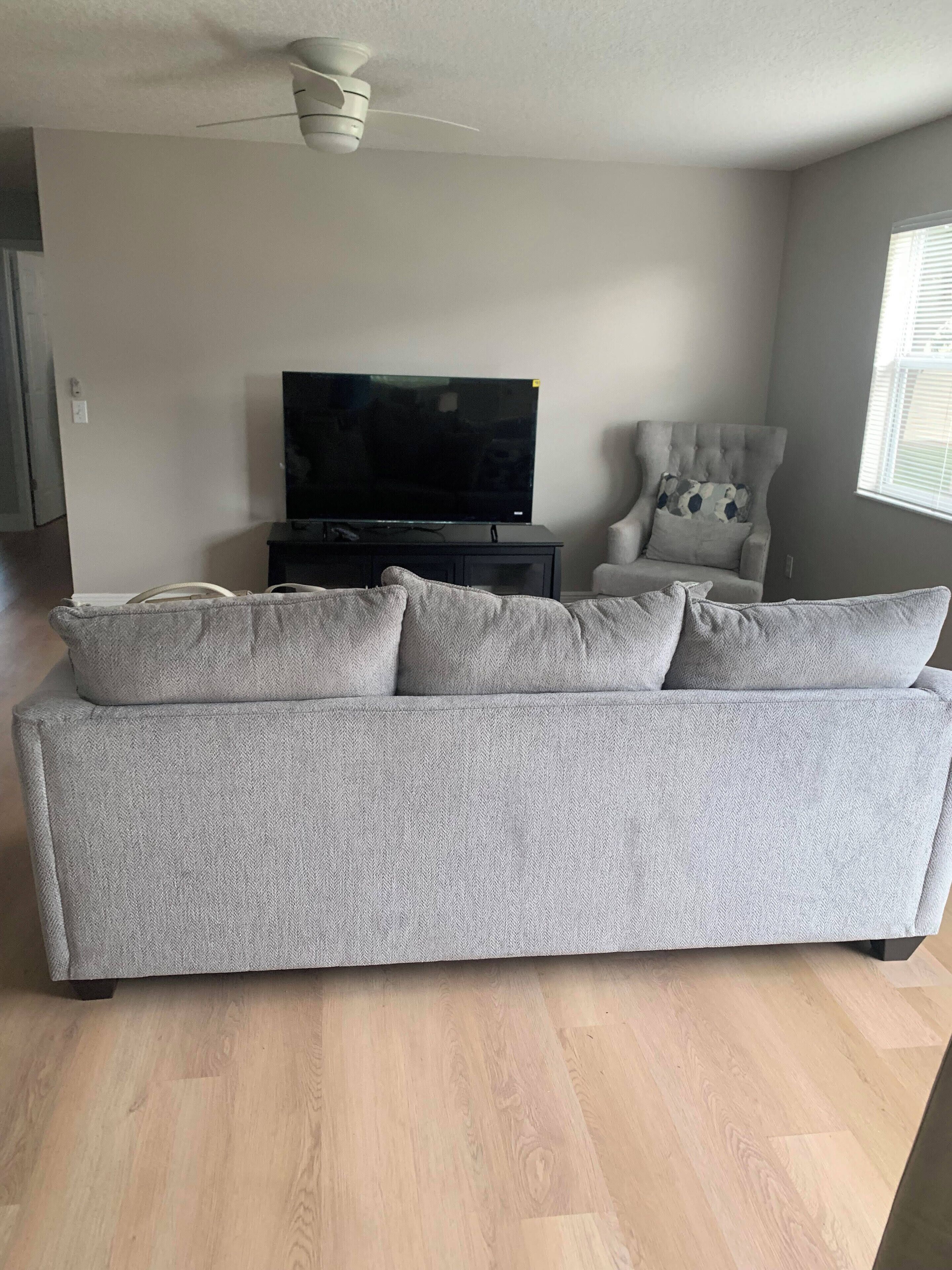 A living room with a grey sofa, a television, a ceiling fan, and a window with blinds.