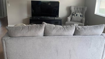 A living room with a grey sofa, a television, a ceiling fan, and a window with blinds.