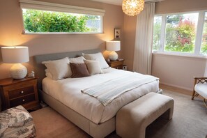 A bedroom with a large bed, two bedside lamps, a nightstand, a chair, and a window with a view of greenery.