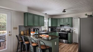 A modern kitchen with green cabinets, a central island with bar stools, a stainless steel refrigerator, and a wooden floor.