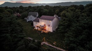 A two-story house with a deck and outdoor lighting, surrounded by dense trees and hills in the background.