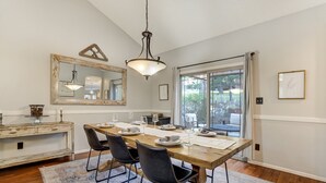 Un coin repas avec une table en bois dressée pour quatre personnes, des chaises noires, un lustre, un miroir et une vue sur l’extérieur grâce à une porte vitrée.