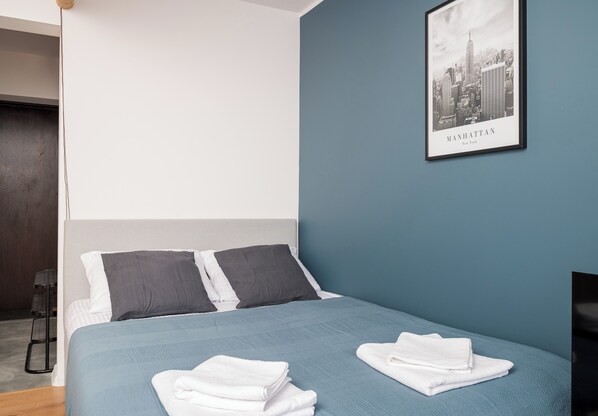 A neatly made bed with a blue bedspread and two pillows, a framed cityscape picture on the wall, and a folded white towel on the bed.