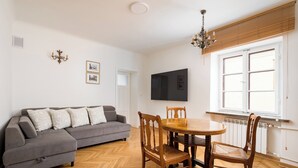 Un salon avec un canapé gris, une table à manger en bois et des chaises, un téléviseur fixé au mur et une fenêtre avec des stores.