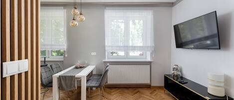 A modern living room with a dining area, a TV mounted on the wall, and a radiator under the windows.