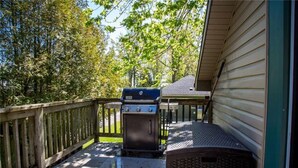 A covered outdoor area with a grill, a wooden deck, and a house in the background. - Cozy 3-Bedroom Beach Cottage (Turkey Point)