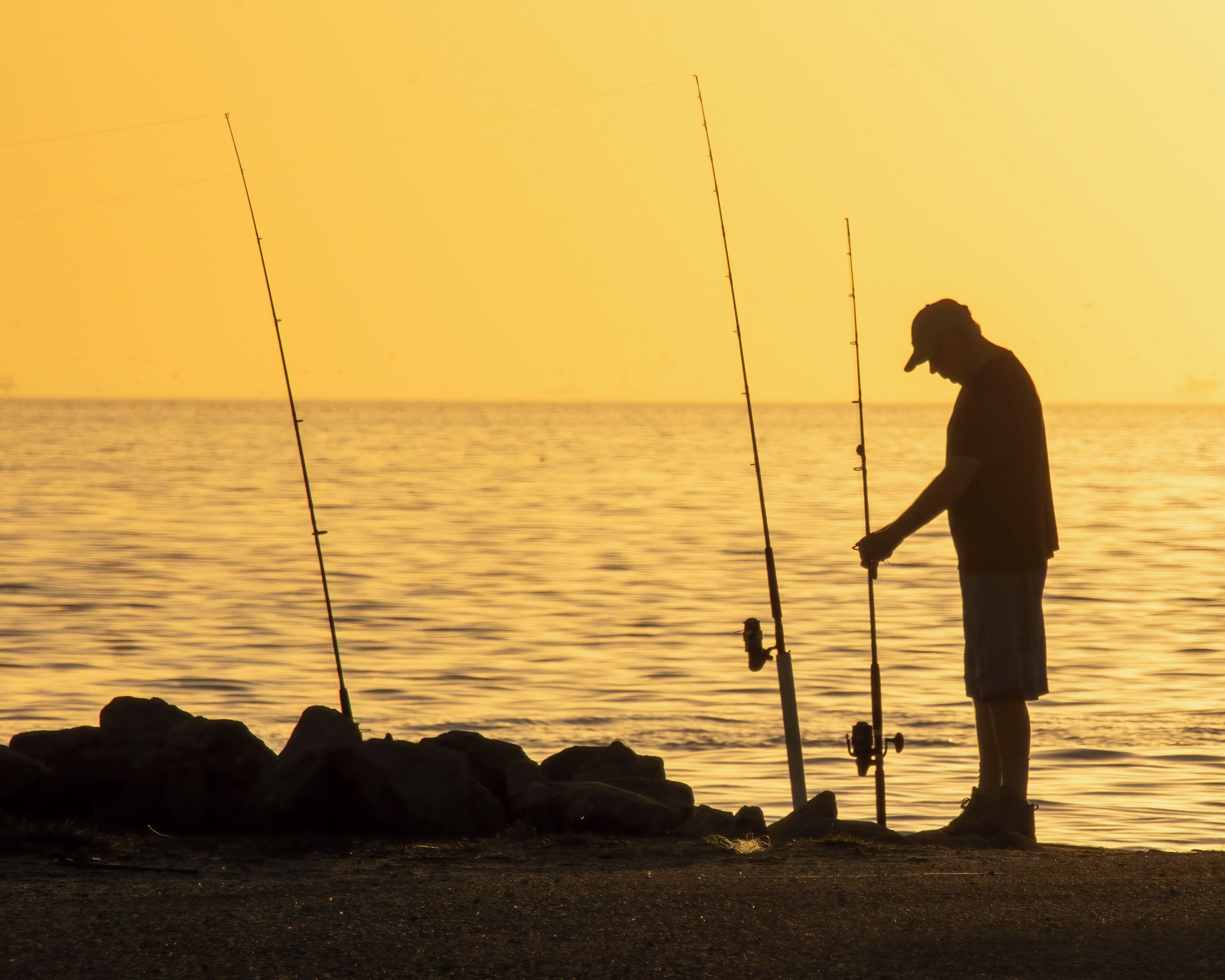 A person fishing at sunset with fishing rods and a boat in the background.