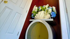 Een kamer met een witte deur, een houten wand, een decoratieve plafondlamp, een bloemstuk en een grote ronde spiegel.
