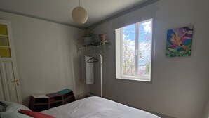 Une chambre à coucher avec un lit, des oreillers, une plante suspendue, une fenêtre et un tableau au mur.