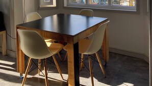 Un espace repas comprenant une grande table et quatre chaises, un luminaire suspendu et une vue sur le paysage urbain grâce à de grandes fenêtres.