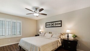 Une chambre à coucher comprenant un lit, deux tables de chevet, un ventilateur de plafond, une fenêtre avec des stores et un élément décoratif fixé au mur.