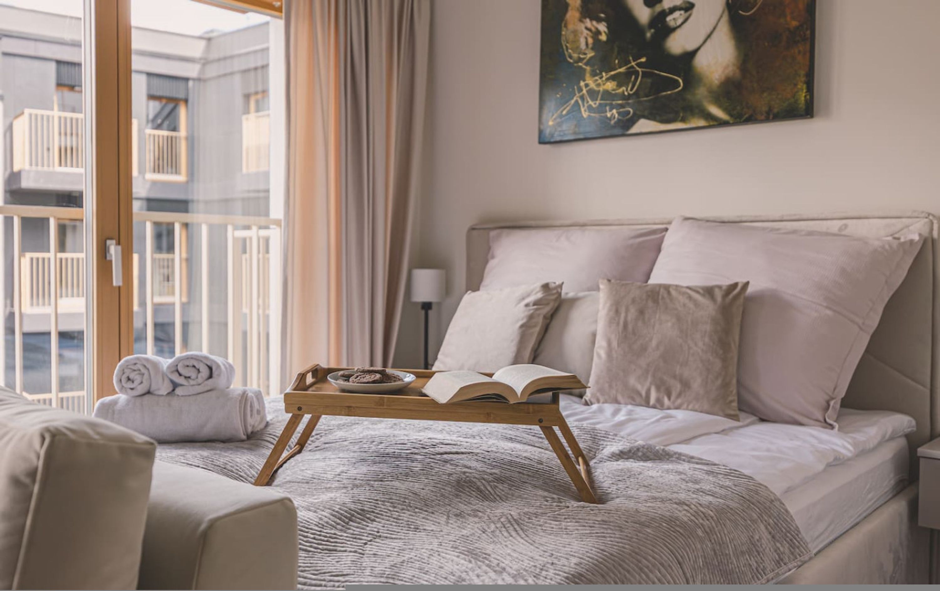 Een hotelkamer met een bed, een nachtkastje met een dienblad met koekjes en een boek, een schilderij aan de muur en uitzicht op het balkon.