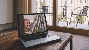 Un ordinateur portable est posé sur une table en bois, et par la fenêtre, on aperçoit un bâtiment et des chaises d’extérieur.