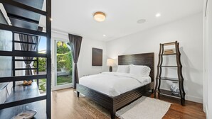A modern bedroom with a large bed, a wooden headboard, a bedside table with a lamp, and a view of the outdoors.