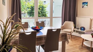 A dining area with a table, chairs, and a bowl of fruit. There are curtains, a radiator, and a plant. - Lovely apartment for 4 people with WIFI, TV and balcony (Wolgast)