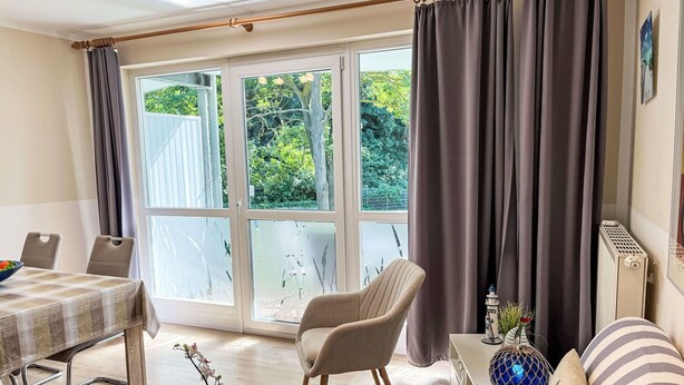 A room with a dining table, a sofa, a chair, and a small table with a plant. There are large windows with curtains, and a radiator is visible on the wall. - Lovely apartment for 4 people with WIFI, TV and balcony (Wolgast)