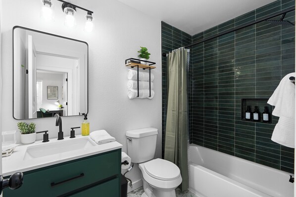 A modern bathroom with a white sink, black faucet, green cabinet, white toilet, and a shower with green tiles.