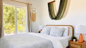 Ein Schlafzimmer mit einem großen Bett, zwei Nachttischen mit Lampen, einem Fenster mit Vorhängen und einem Wandbehang aus Makramee.