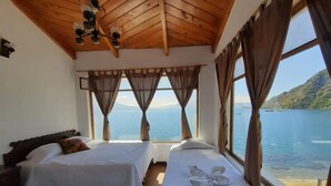 Una habitación con dos camas, amplias ventanas con cortinas, techo de madera y vistas a un lago y montañas.
