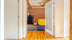 A hallway with wooden flooring, a brick wall, and a door leading to a room with a bed and a dresser.