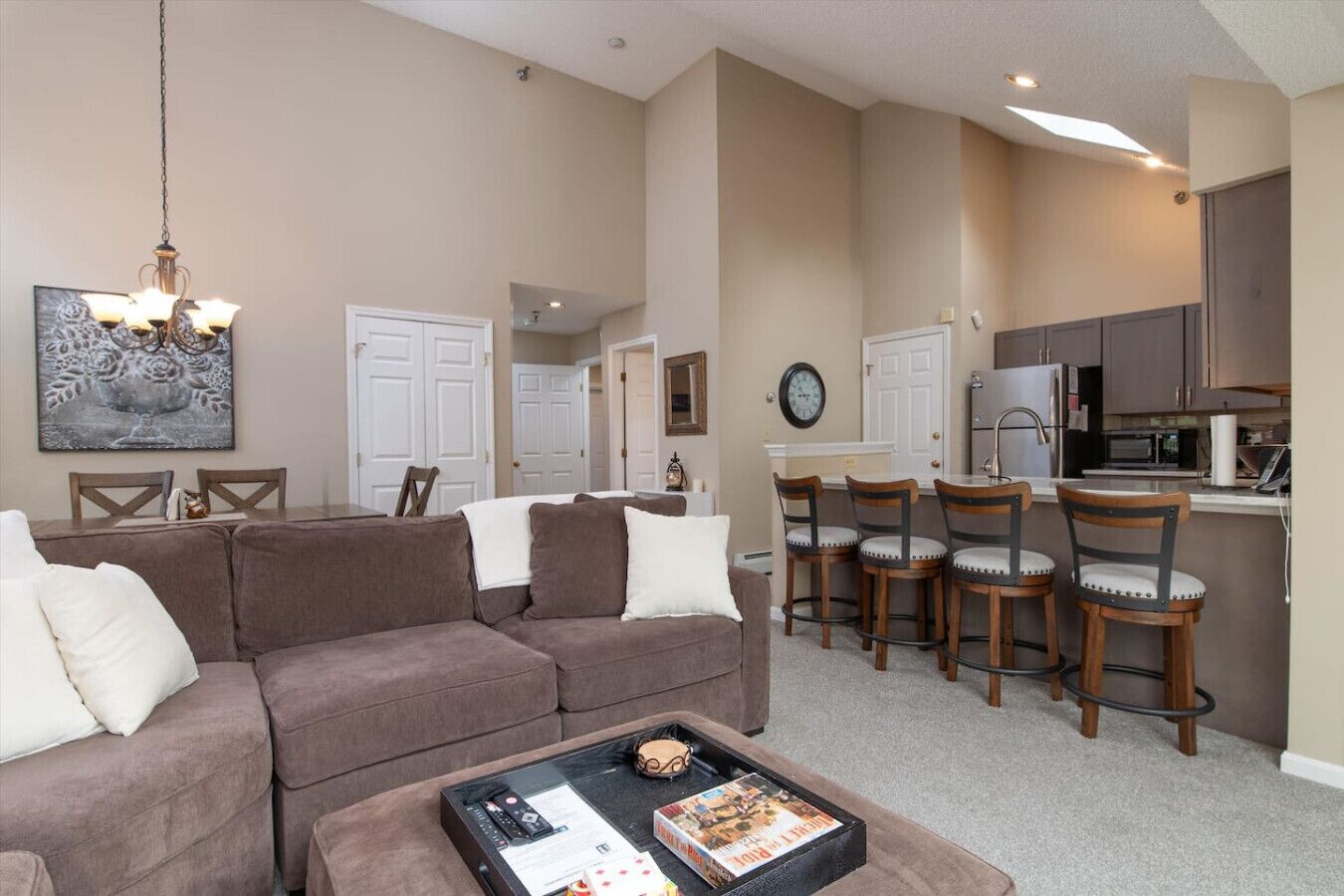 A living room with a brown sofa, a coffee table with magazines, and a dining area with wooden chairs and a kitchen in the background.