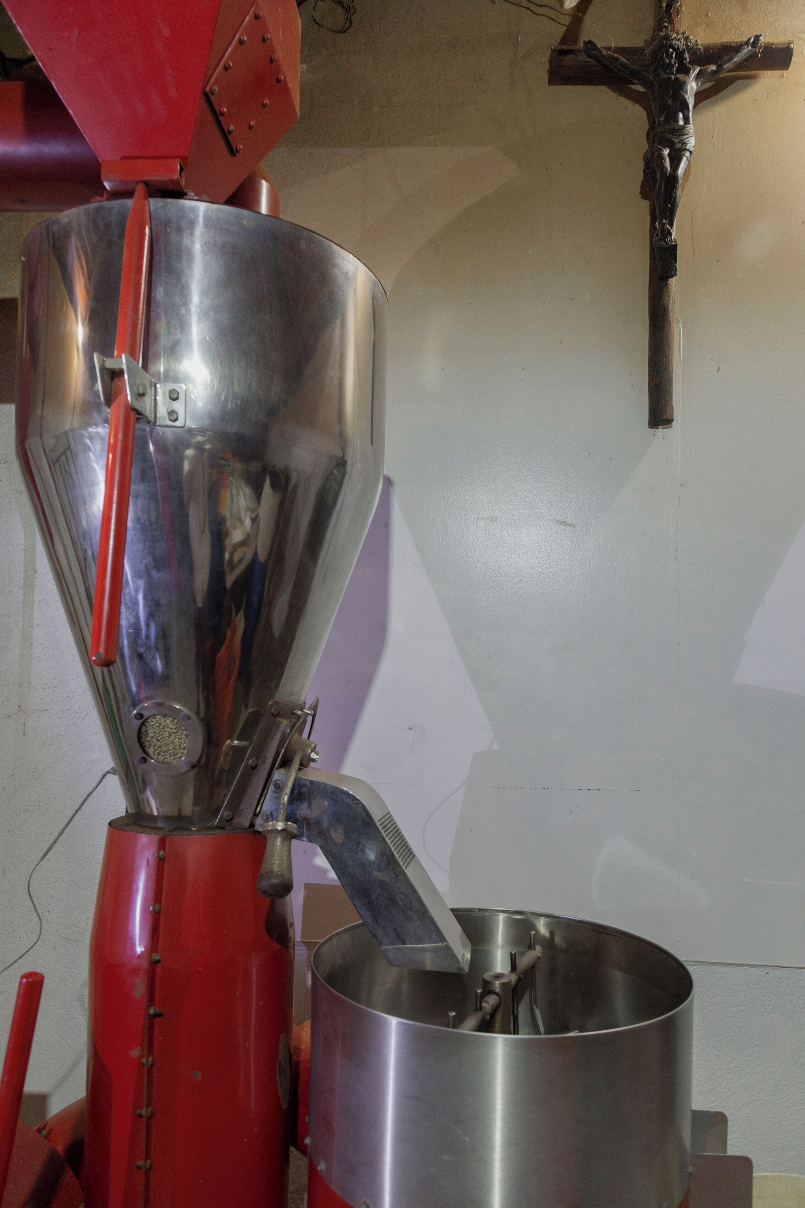Una cafetera con un embudo metálico y una jarra, una máquina roja, y una cruz en la pared.