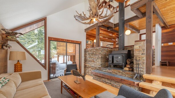 A cozy living room with a stone fireplace, wooden beams, and a large window overlooking a snowy landscape.