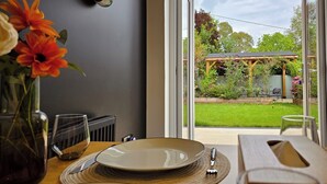 Un comedor con una mesa preparada para dos, un jarrón con flores y vista a un jardín por una puerta de cristal.