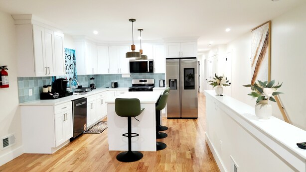 A modern kitchen with white cabinets, a central island with bar stools, a microwave, a refrigerator, and a coffee machine. - Luxury 3BR Retreat Near NYC (Newark)