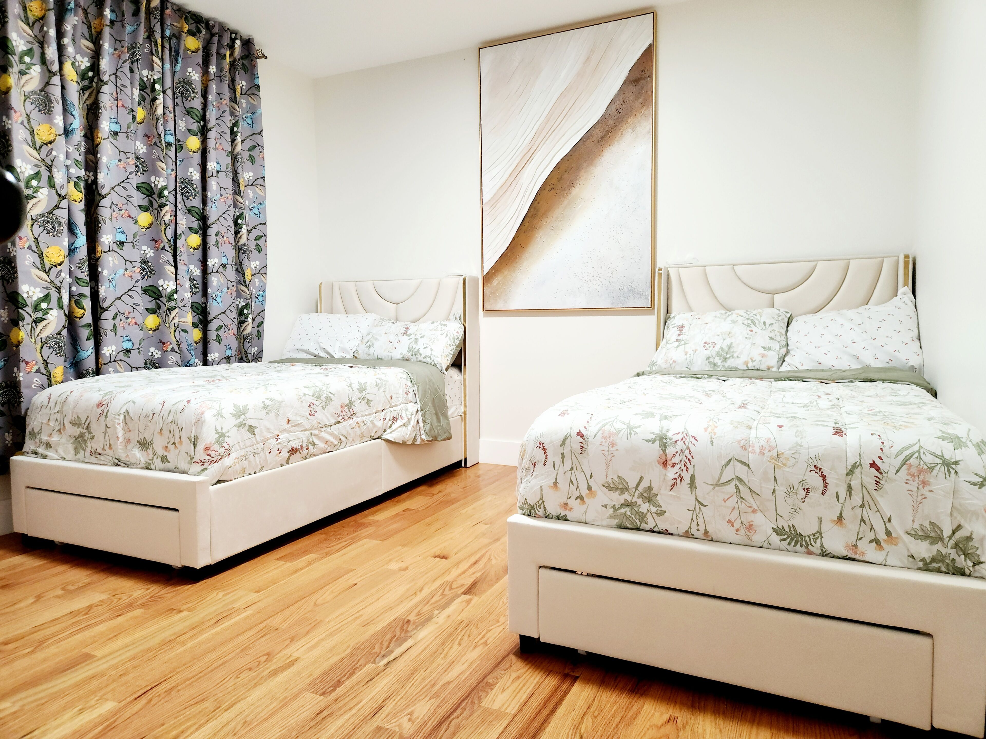 Une chambre dotée de deux lits, chacun recouvert d’une couvre-lit à motifs floraux et agrémenté d’oreillers assortis, avec un parquet et une grande peinture abstraite au mur.