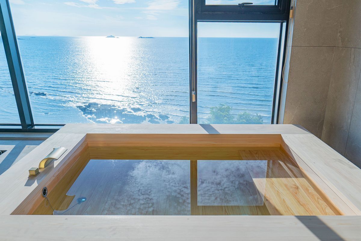 A modern bathroom with a large bathtub, a window with a view of the sea, and a wooden floor.