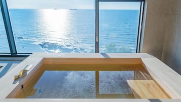 A modern bathroom with a large bathtub, a window with a view of the sea, and a wooden floor.