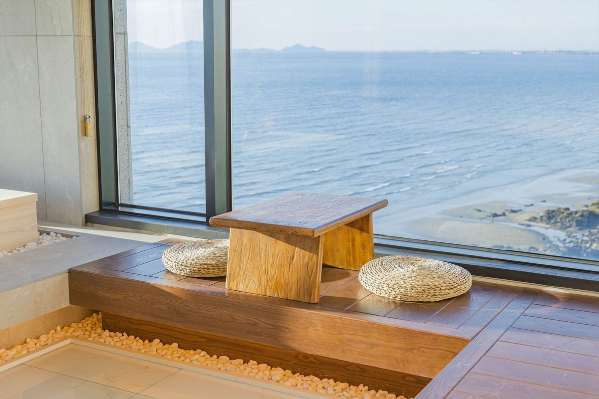 A wooden bench with woven baskets, a stone ledge, and a view of the ocean through a large window.