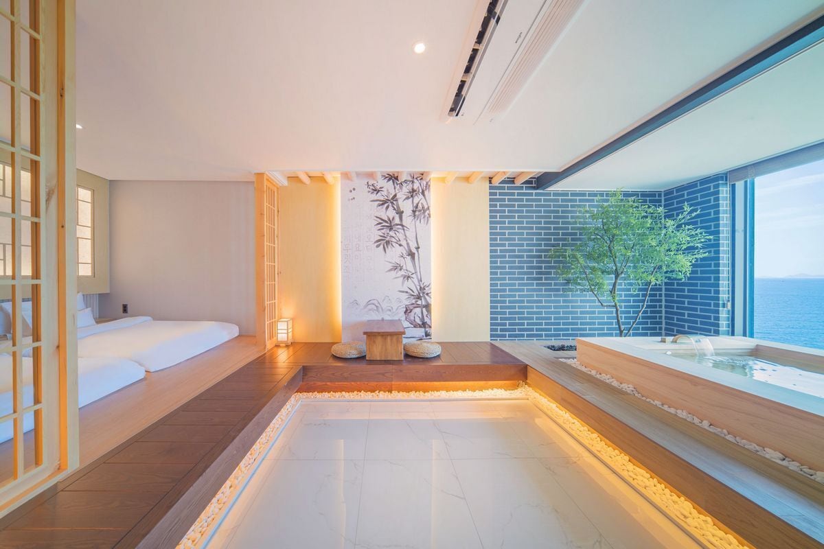 A modern spa room with a wooden floor, a seating area with cushions, a small wooden table, a wall with a bamboo pattern, and a large window overlooking the sea.