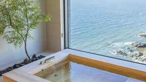 Un área de spa con una bañera de madera, un pequeño árbol y vista al océano a través de un ventanal grande.