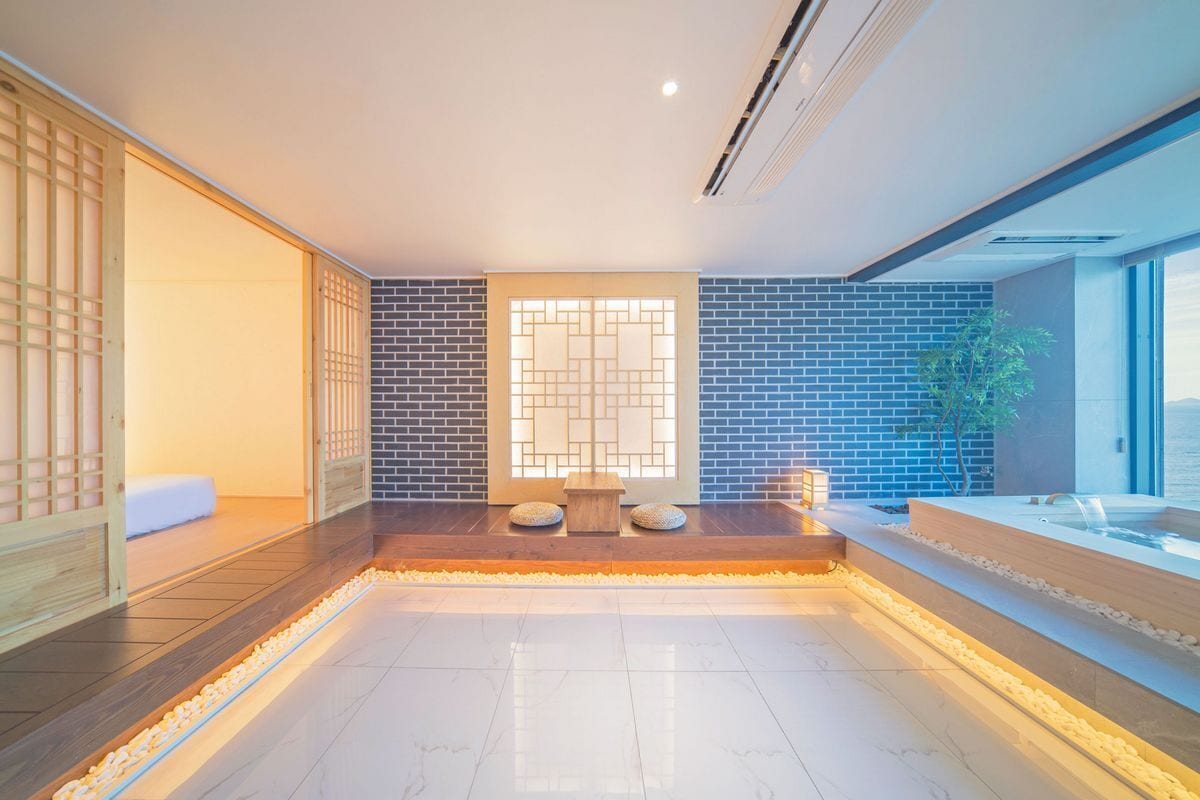 A modern spa room with a large, tiled floor, a wooden bench, a small table, and a water feature.