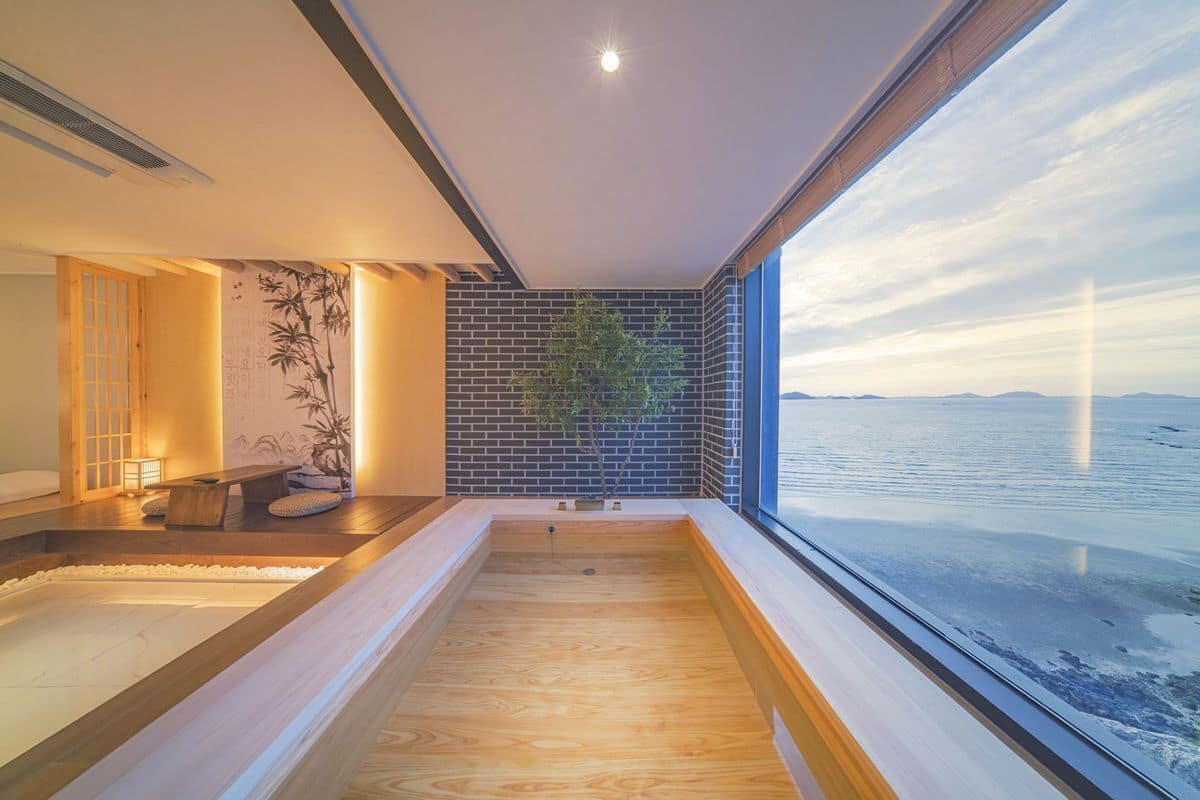 Un spa moderno con suelo de madera, una pequeña zona de estar, una planta en la pared y un ventanal grande con vistas a un cuerpo de agua.