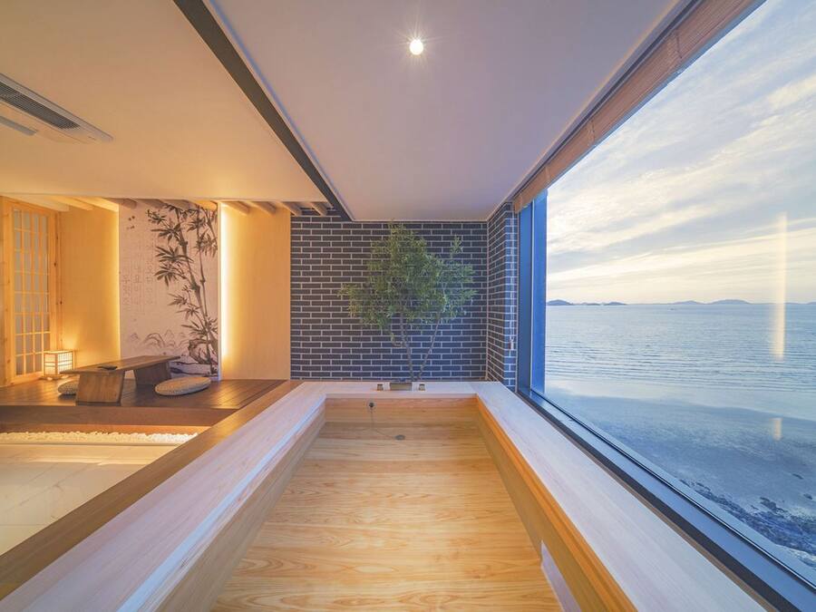 Ein moderner Wellnessraum mit Holzboden, einer kleinen Sitzecke, einer Wandpflanze und einem großen Fenster mit Blick auf ein Gewässer.