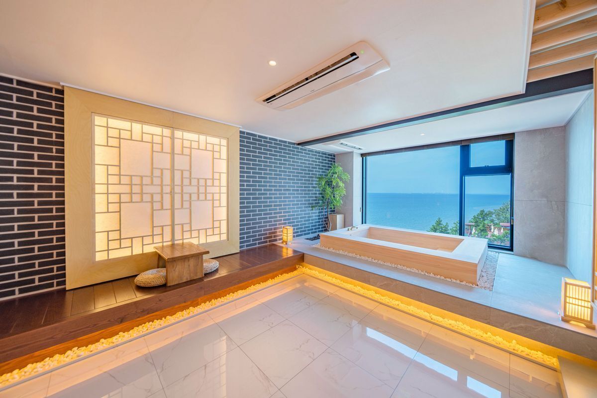 A modern bathroom with a large bathtub, a wooden bench, and a view of the sea.