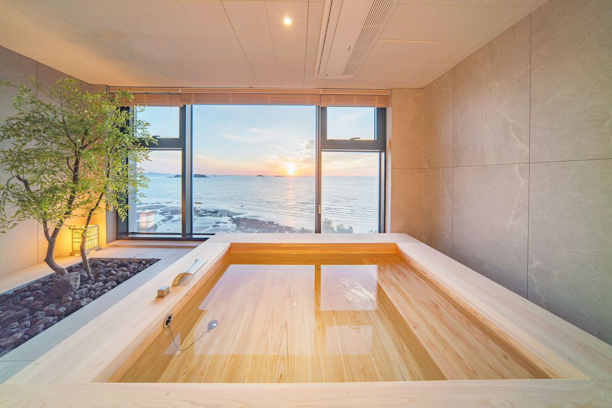Un baño moderno con bañera de madera, un pequeño árbol y vista al mar al atardecer.