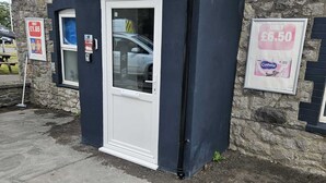 A small, dark-colored building with a white door and a window. There are two signs on the wall: one indicating a price of £1.65 and another showing £6.50. - 5 beds/Spacious 4-Bedroom Retreat in the Heart of Churchill  with Parking (North Somerset)