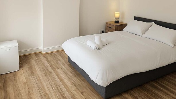 A modern bedroom with a large bed, a bedside table with a lamp, and a small white appliance. - 5 beds/Spacious 4-Bedroom Retreat in the Heart of Churchill  with Parking (North Somerset)