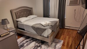 Ein ordentlich bezogenes Bett mit weißen Leinen und einer grauen Decke, eine Nachttischlampe und ein Teppich mit Marmormuster.