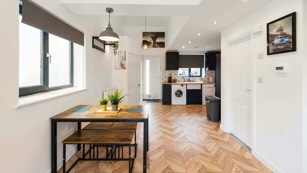 A modern kitchen with a built-in washing machine, a dining area with a wooden table and bench, and a hallway with a herringbone floor pattern. - Chic Urban Haven with Double Bed House (Bristol City)