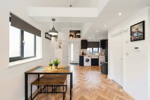 A modern kitchen with a built-in washing machine, a dining area with a wooden table and bench, and a hallway with a herringbone floor pattern. - Chic Urban Haven with Double Bed House (Bristol City)