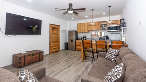 A living room with a sofa, a wooden chest, and a TV on the wall. There is a kitchen area with wooden cabinets and a dining table with chairs.